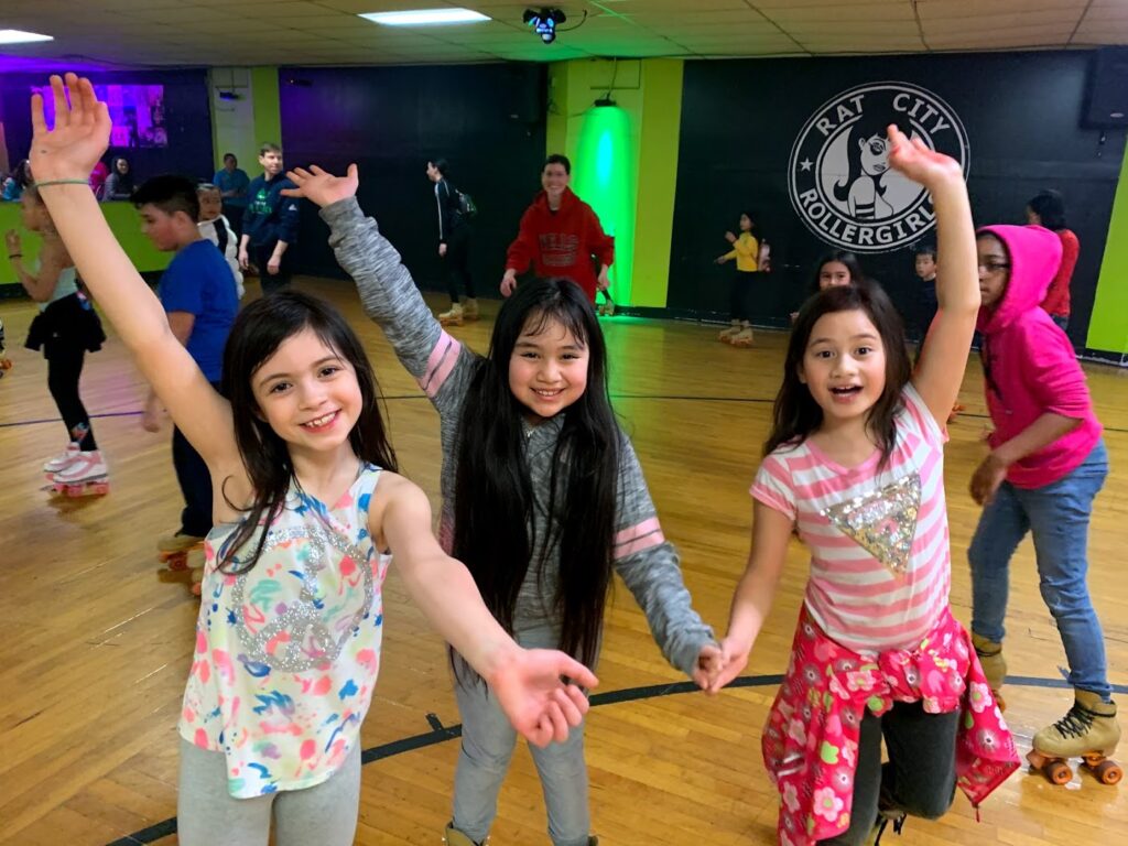 Three students in a wooden roller skating rink raise one arm and lower the other like an alligator clap